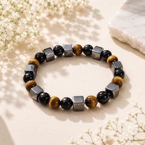 Könnte beinhalten: Ein Perlenarmband mit abwechselnd runden schwarzen Perlen, runden braunen Tigeraugen-Perlen und glatten metallisch grauen würfelförmigen Perlen, die in einem sich wiederholenden Muster um das kreisförmige Band angeordnet sind.