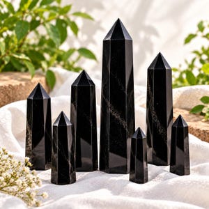 Può includere: Una collezione di sette torri in pietra nera lucida con cime esagonali appuntite. Ogni torre presenta una superficie liscia e riflettente con sottili venature naturali che corrono in diagonale sul materiale scuro e opaco dei cristalli.