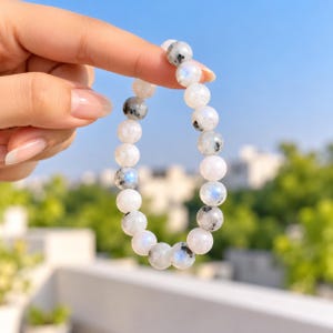 May include: A stretch bracelet composed of round, polished beads in shades of white, translucent gray, and black. Several beads feature a distinct blue iridescent sheen, while others display natural dark inclusions and speckles.