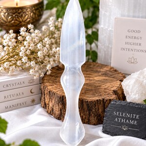 May include: A translucent white selenite athame with a pointed blade and a contoured handle. The object features a smooth, polished surface with natural crystalline striations. A small black sign with white text reads SELENITE ATHAME.
