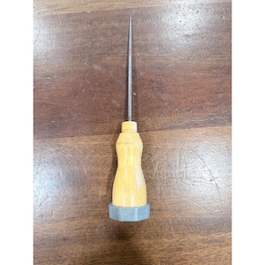 May include: A hand tool featuring a long, thin, pointed metal spike attached to a smooth, light-colored wooden handle. The base of the handle is fitted with a hexagonal gray metal nut, providing a sturdy grip for the pointed metal shaft.