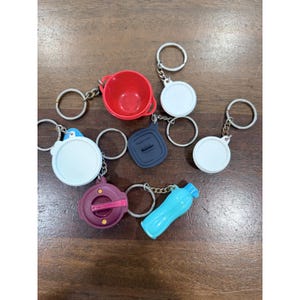 May include: A collection of miniature Tupperware-style keychains featuring a small red bowl, a bright blue water bottle, a dark purple container lid, a dark blue square lid, and three white circular lids, all attached to silver metal key rings.