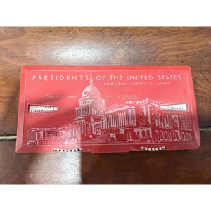 Op de afbeelding: Een rechthoekig rood plastic educatief apparaat met de titel PRESIDENTS OF THE UNITED STATES and their terms in office. Het bevat een witte afbeelding van het Amerikaanse Capitool, met kleine vensters die tekst tonen en twee witte draaiwielen.
