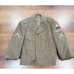 May include: An olive drab wool military jacket featuring two front chest pockets, a pointed collar, and long sleeves. The left sleeve displays a triangular patch with yellow, red, and blue sections, and both sleeves feature yellow chevron rank insignia.