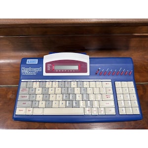 May include: A blue VTech Keyboard Wizard electronic learning device featuring a QWERTY keyboard with white and grey keys, a numeric keypad, and a central LCD screen with a white and maroon housing. The unit includes various function buttons.
