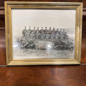 May include: A black and white photograph in a gold-toned frame depicting a large group of soldiers in uniform. A sign in the center reads 3RD PLATOON COMPANY C 3052 ENGINEER COMBAT BATTALION GERMANY, 8 MAY 1945. The soldiers are posed in rows on a field.
