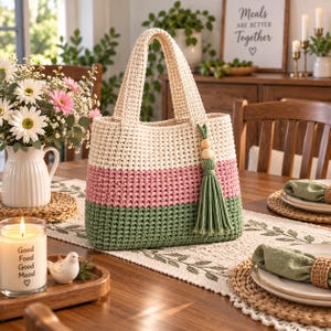 Pode incluir: Uma bolsa tote de crochê com listras horizontais em creme, rosa antigo e verde sálvia. A bolsa possui duas alças resistentes e um detalhe de borla decorativa preso em um dos lados, com duas contas de madeira clara em um cordão.