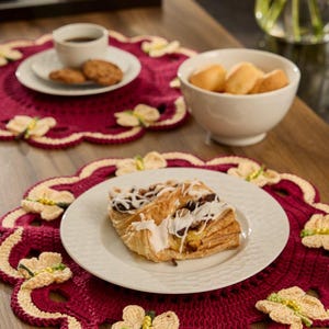Pode incluir: Um lugar americano redondo de crochê em vermelho escuro com uma borda festonada na cor creme, decorado com vários pequenos motivos de borboletas de crochê na cor creme. Cada borboleta é acentuada por uma fileira vertical de pequenas contas amarelas.