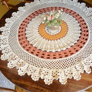 Puede incluir: Un mantel redondo de ganchillo multicolor con anillos concéntricos en tonos crema, amarillo mostaza y terracota. El diseño incluye intrincados patrones calados, un borde festoneado y un arreglo floral central en un florero de vidrio.