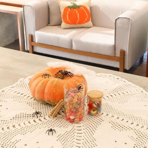 May include: A round white crocheted doily featuring ghost patterns, topped with an orange pumpkin, two glass jars filled with colorful candies, and several small black plastic spider figurines. A thin layer of white faux spiderweb is present.