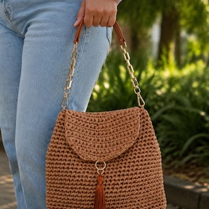 Pode incluir: Uma bolsa de mão em crochê castanha com um padrão de pontos texturizados, uma aba dobrável e um fecho central de anel dourado com uma borla castanha a condizer. A bolsa inclui uma alça de corrente dourada com detalhes em imitação de pele.