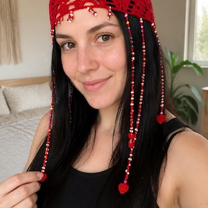 Pode incluir: Um acessório de cabeça em crochê vermelho brilhante com um padrão rendado intrincado. A peça é adornada com vários fios pendentes de contas multicoloridas e pequenos corações de crochê vermelhos nas pontas de cada cordão.