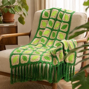 May include: A crochet throw blanket featuring a repeating square pattern with bright lime green leaf motifs centered on cream-colored backgrounds, framed by dark green borders and finished with long dark green fringe along the bottom edge.