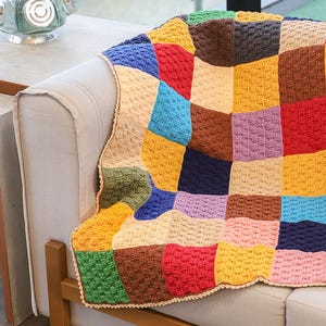Pode incluir: Uma manta de crochê feita à mão com um design de patchwork de blocos quadrados em várias cores, incluindo vermelho, amarelo, azul, verde, marrom, rosa e roxo. A manta tem um padrão de ponto texturizado e uma borda festonada bege clara.