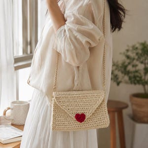 Pode incluir: Uma bolsa transversal de crochê bege em estilo envelope, com uma pequena aplicação de coração vermelho texturizado na aba frontal. A bolsa é suspensa por uma fina alça de corrente de metal dourado, exibindo uma construção detalhada.