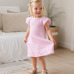 Pode incluir: Um vestido de crochê rosa claro com mangas curtas com babados, decote redondo e um padrão de pontos texturizados em todo o corpete, finalizado com uma borda decorativa vazada ao longo da bainha, usado com sandálias bege de tiras.