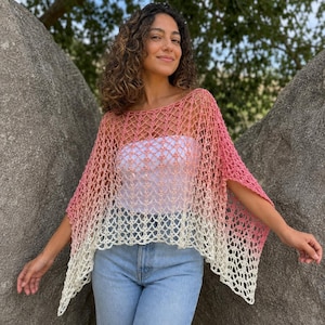 Pode incluir: Um poncho de crochê com um padrão de rede aberto e um gradiente de cor que transita do rosa escuro no topo para um tom de rosa mais claro no meio e branco na bainha inferior. A peça tem um decote largo e mangas curtas.