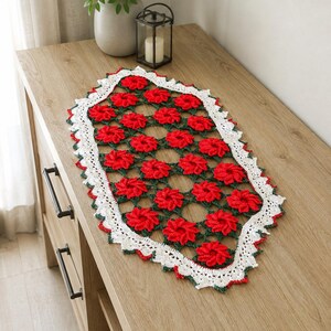 Pode incluir: Um caminho de mesa oval de crochê com um padrão repetitivo de motivos florais vermelho-brilhantes conectados por fio verde-escuro, finalizado com uma borda decorativa branca festonada com pequenos detalhes vermelhos e verdes.