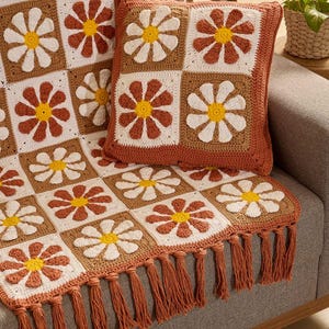 Pode incluir: Uma manta de crochê e uma almofada quadrada a condizer com um motivo repetitivo de margaridas. O design consiste em pétalas brancas e cor de ferrugem com centros amarelos em blocos quadrados. A manta tem franjas compridas cor de ferrugem.