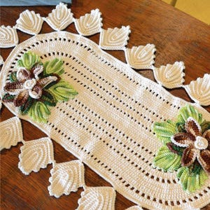 Pode incluir: Um caminho de mesa de crochê de cor creme com formato oval e uma borda decorativa de motivos em forma de folha. Dois grandes adornos florais em tons de castanho, branco e verde estão fixados nas extremidades do caminho, adicionando textura.