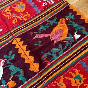 May include: A colorful woven rug with a geometric pattern and a bird design. The rug is made of wool and has a variety of colors, including red, orange, yellow, green, and blue.