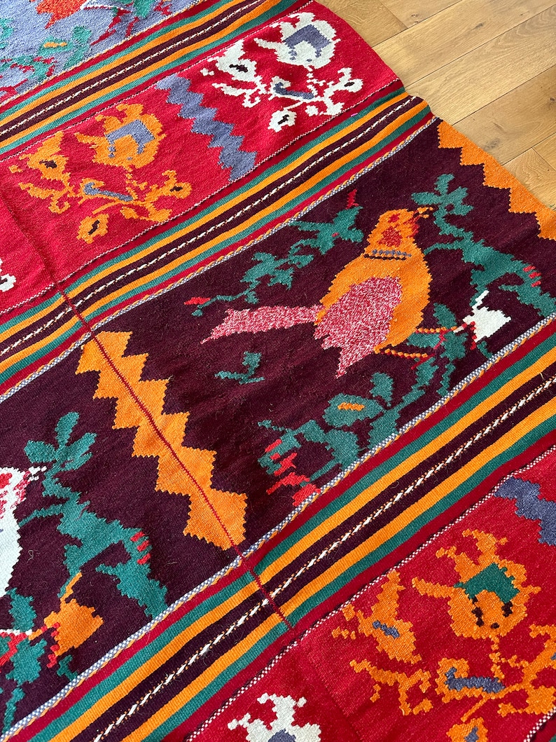 May include: A colorful woven rug with a geometric pattern and a bird design. The rug is made of wool and has a variety of colors, including red, orange, yellow, green, and blue.