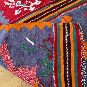 May include: A colorful woven rug with a geometric pattern in shades of red, orange, yellow, green, blue, and white. The rug has a fringed edge and is folded in half.
