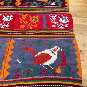 May include: A colorful woven rug with a geometric pattern and a bird design. The rug has a fringe border and is made of wool.