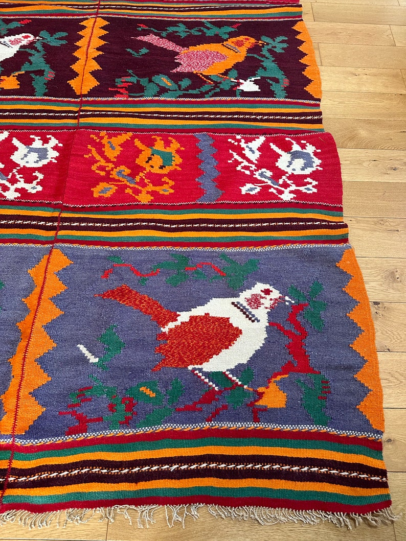 May include: A colorful woven rug with a geometric pattern and a bird design. The rug has a fringe border and is made of wool.