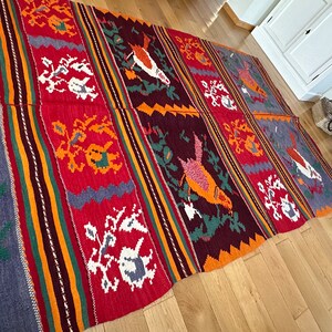 May include: A colorful woven rug with a geometric pattern featuring birds and floral motifs. The rug is made of wool and has a traditional design.