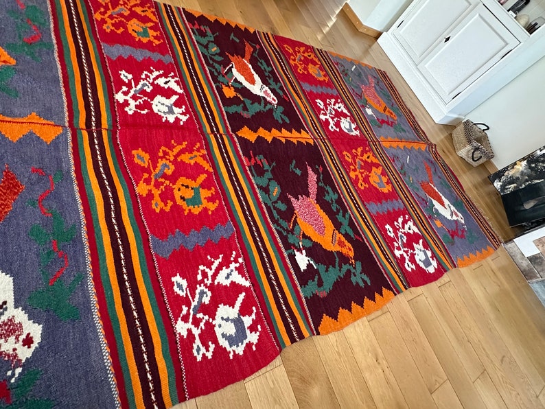 May include: A colorful woven rug with a geometric pattern featuring birds and floral motifs. The rug is made of wool and has a traditional design.