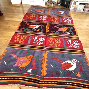 May include: A colorful woven rug with a repeating pattern of birds and floral designs. The rug is made of wool and has a fringed edge.