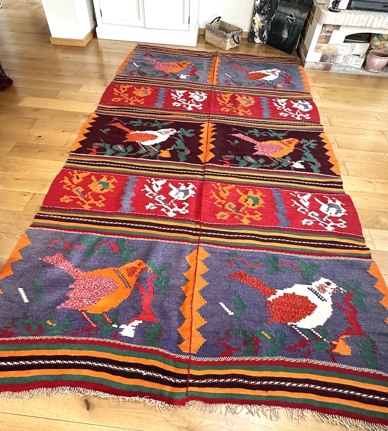 May include: A colorful woven rug with a repeating pattern of birds and floral designs. The rug is made of wool and has a fringed edge.