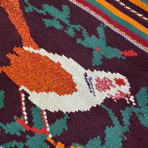 May include: A close-up of a woven rug with a colorful design featuring a bird in the center. The bird is white with orange and red accents, and is surrounded by green and red leaves. The rug has a dark brown background with stripes of orange, yellow, green, and red.