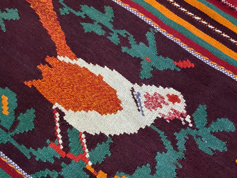 May include: A close-up of a woven rug with a colorful design featuring a bird in the center. The bird is white with orange and red accents, and is surrounded by green and red leaves. The rug has a dark brown background with stripes of orange, yellow, green, and red.