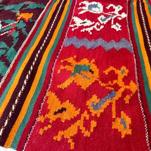 May include: A close-up of a colorful woven rug with intricate patterns in shades of red, orange, green, blue, and white. The rug has a traditional design with geometric shapes and floral motifs.