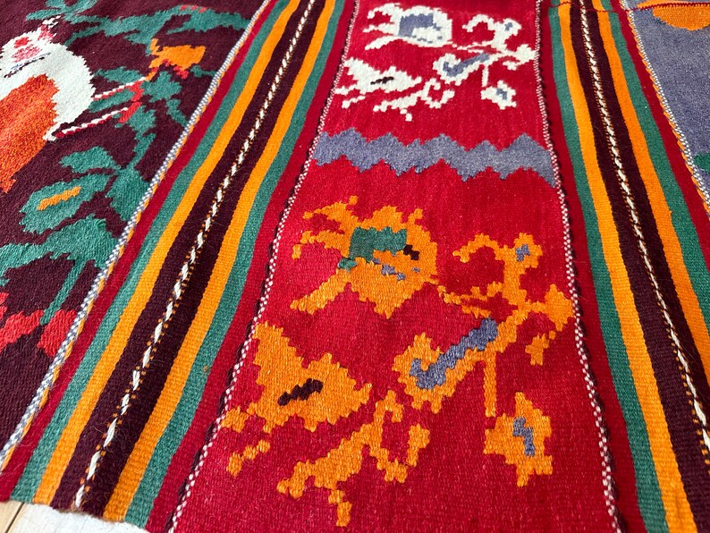 May include: A close-up of a colorful woven rug with intricate patterns in shades of red, orange, green, blue, and white. The rug has a traditional design with geometric shapes and floral motifs.