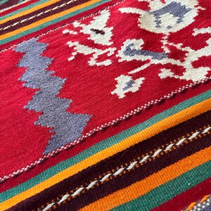 May include: A close-up of a colorful woven rug with a geometric pattern. The rug features stripes of red, orange, green, and brown, with a white floral design in the center.