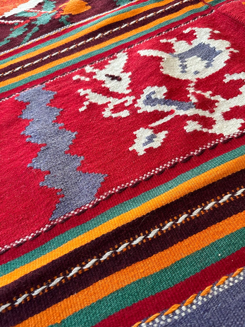 May include: A close-up of a colorful woven rug with a geometric pattern. The rug features stripes of red, orange, green, and brown, with a white floral design in the center.