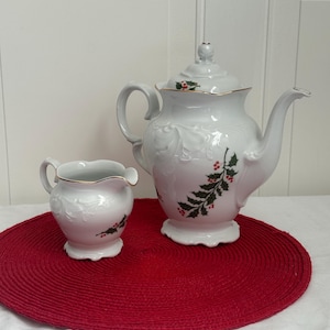 May include: A white porcelain tea set with a teapot and creamer. The set features embossed floral designs and is decorated with holly and red berries. The teapot has a gold trim and a lid. The set is on a red circular placemat.