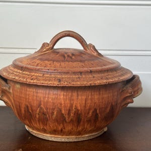 Puede incluir: Una cacerola de cerámica marrón con tapa y dos asas. La cacerola tiene un aspecto rústico y artesanal con un esmalte moteado.