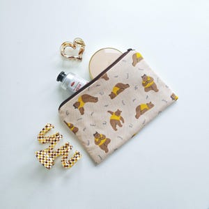 Op de afbeelding: Een beige etui met rits voorzien van een herhalend patroon van bruine beren in gele shirts, vergezeld door een hartvormige schildpadkleurige haarklem, een ronde compactspiegel, een kleine zilveren tube en een geblokte bruin-witte haarklem.