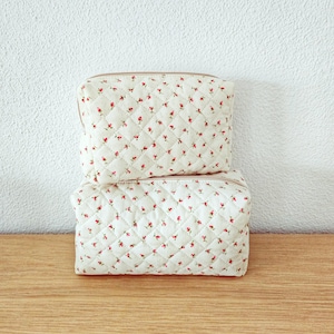 Peut inclure: Deux trousses rectangulaires matelassées empilées verticalement, présentant une base en tissu blanc cassé avec un motif répétitif de minuscules boutons de rose rouges et de feuilles vertes. La texture matelassée forme un quadrillage en losanges.