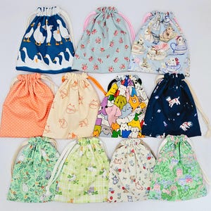 Peut inclure: Une collection de onze pochettes à cordon en tissu présentant divers motifs, notamment des oies blanches sur fond bleu, des imprimés floraux, des lapins dans des tasses, des pois orange, des chats au trait, des chiens colorés et des motifs de jardin.