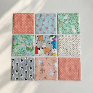 Op de afbeelding: Een set van negen vierkante stoffen onderzetters in een raster. De patronen bevatten koraalkleurige stippen, lichtblauwe bloemen, groen met witte konijnen, een tuinscène, kleurrijke abstracte diervormen, een bloemenrooster, gestreept klaverblad en perzikkleurige bloemen.