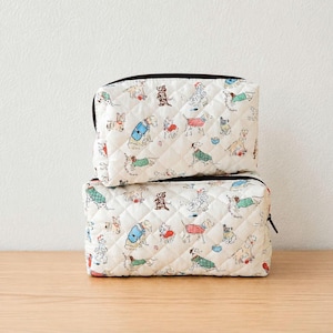 Puede incluir: Dos estuches acolchados con cremallera apilados verticalmente, con una base de tela color blanco hueso y un patrón repetitivo de varias razas de perros con suéteres y accesorios coloridos. El acolchado crea un diseño de costuras en rombos.