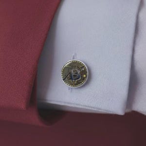 May include: A circular cufflink featuring a metallic gold and black Bitcoin symbol with an upward-trending arrow design. The cufflink is fastened through the buttonhole of a crisp white shirt cuff, which is partially covered by a dark red jacket sleeve.