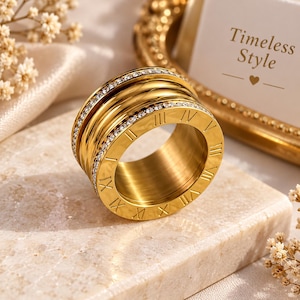 May include: A wide gold-toned ring featuring engraved Roman numerals around the outer edge and two rows of small, clear, sparkling stones bordering the center section. The band has a polished metallic finish and a thick, cylindrical profile.