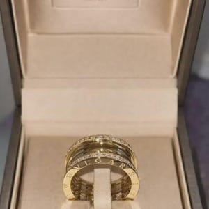 Può includere: Un anello Bvlgari in oro con un design a più fasce e diamanti pavé incastonati lungo i bordi superiore e inferiore. La parola BVLGARI è incisa sulle superfici piatte in oro. L'anello è esposto all'interno di una scatola di marca chiara.