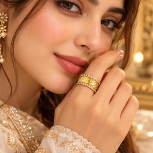 Peut inclure: Une large bague en or avec une section centrale texturée de côtes diagonales, bordée par deux rangées de petites pierres rondes et transparentes. La bague est portée au doigt, accompagnée d'une boucle d'oreille en or assortie à motif floral.